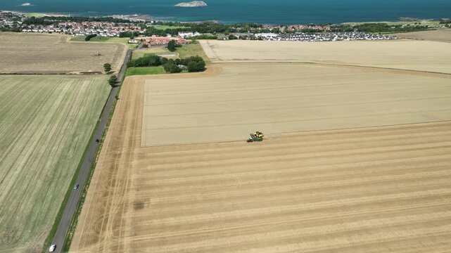 Drone shot of a yellow combine harvester cutting wheat near North Berwick with seaside houses a holiday park and the Firth of Forth in the background
