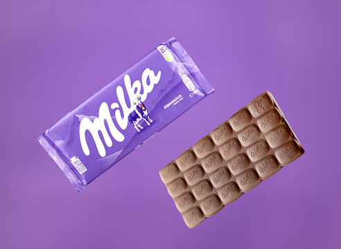 Studio still life of Milka Alpenmilch Alpine milk chocolate, featuring its purple branded packaging beside an unwrapped bar with embossed Milka lettering and a cow motif, arranged diagonally on a purp