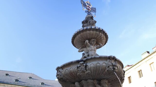 Historic Baroque fountain at Prague Castle features carved stone figures supporting an ornate basin as water gently flows with Gothic cathedral spires rising in the background