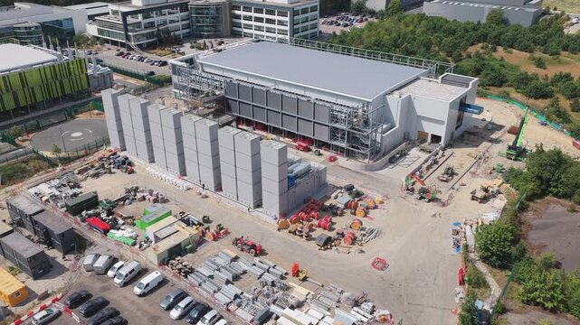 Large data center facility under construction featuring concrete panels and heavy machinery in an industrial setting surrounded by lush green trees and nearby office buildings.