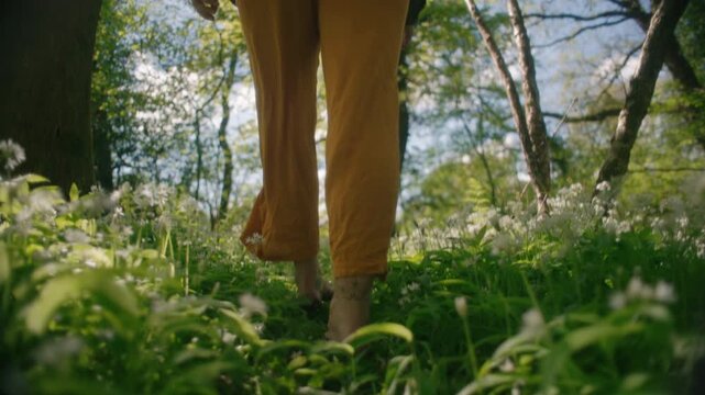 A barefoot woman hops playfully through a carpet of wild garlic flowers in the sunlit woodland.