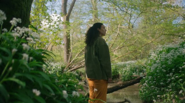 A woman stands near a fallen log over a stream, surrounded by wild garlic flowers in the quiet woodland.