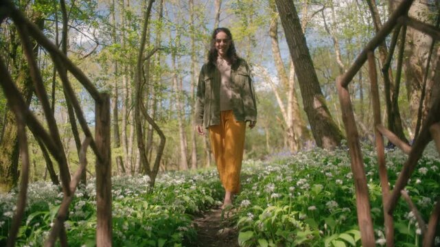 A woman walks along a woodland path flanked by wild garlic flowers and a curved branch archway on a sunny day.