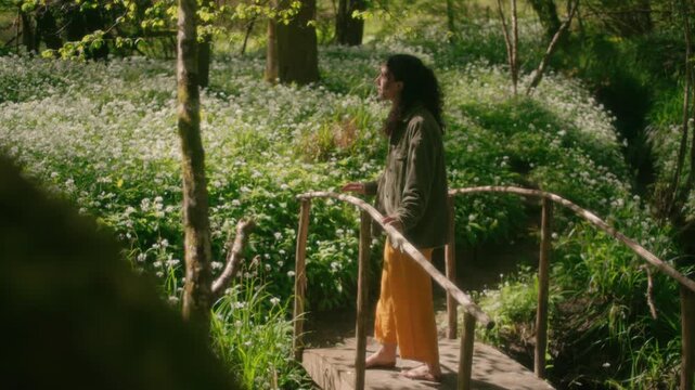 A woman crosses a small rustic wooden bridge over a carpet of wild garlic flowers in the woodland.