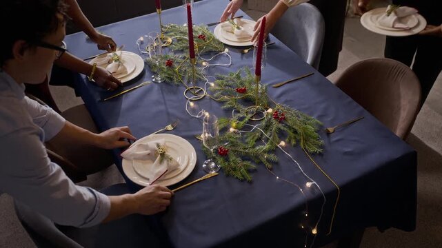 Multiethnic young adult friends setting festive holiday table, placing dinner plates with napkins from top view. Footage suits Christmas celebration commercials and winter holiday promotions