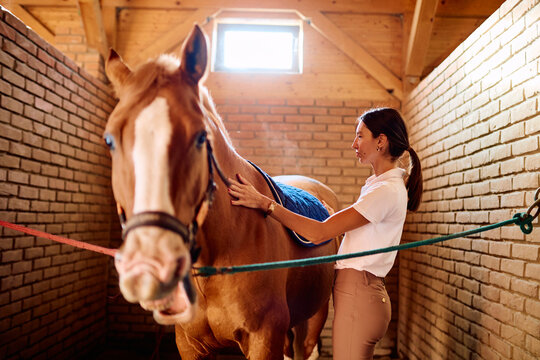 Woman gently petting a brown horse, preparing it for a ride inside a traditional stable with brick walls and wooden beams