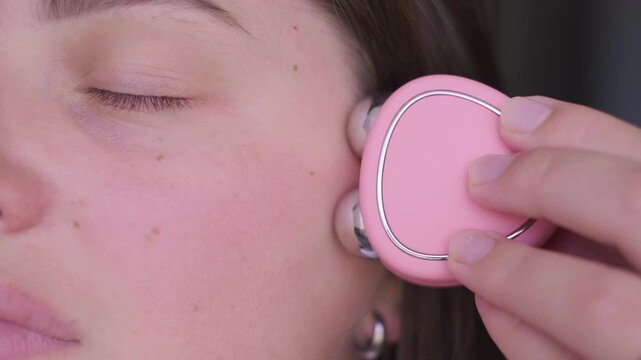 Woman using pink microcurrent device for facial lifting. Close-up on natural skin texture and high-tech beauty gadget. Professional anti-aging skincare routine at home.