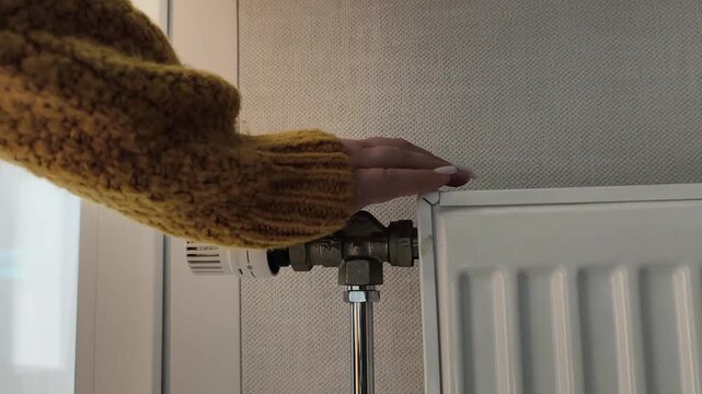 Heating crisis temperature adjustment amidst a heat shortage. A woman in a yellow sweater turns a thermostat dial. Heating season, turning on the heat, cold weather, energy conservation