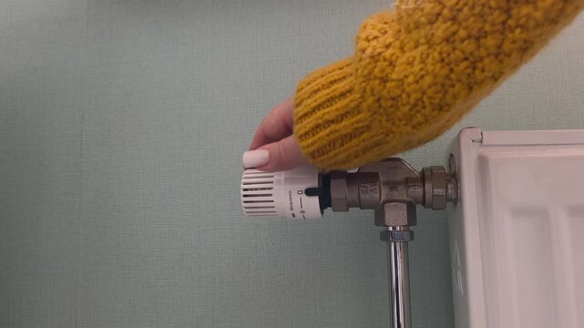 Adjusting the radiator temperature a hand in a yellow sweater adjusts the thermostatic valve. Heating season, turning on the heat, cold time of year
