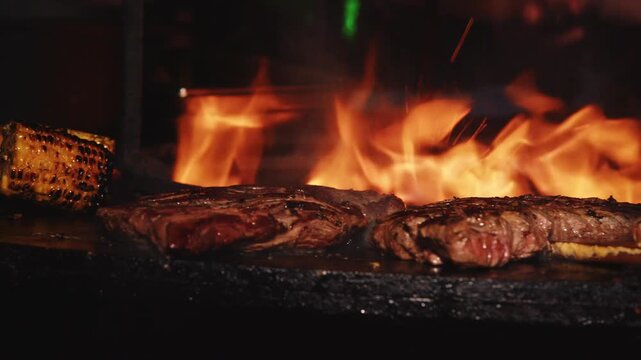 Steak sizzling on a hot griddle next to an open fire, with flames in the background and the meat slowly searing over intense heat.