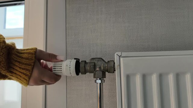Adjusting the radiator temperature a hand in a yellow sweater adjusts the thermostatic valve. Heating season, turning on the heat, cold time of year