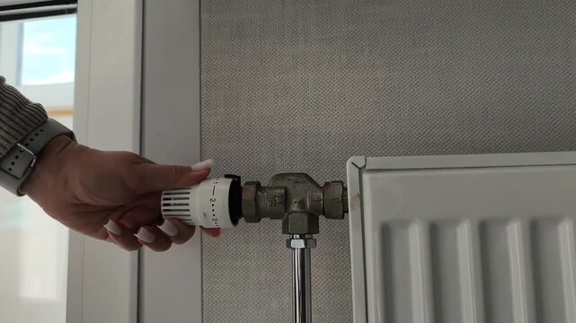Heating crisis temperature adjustment amidst a heat shortage. High cost of electricity. A woman turns a thermostat dial. Heating season, turning on the heat, cold weather, energy saving