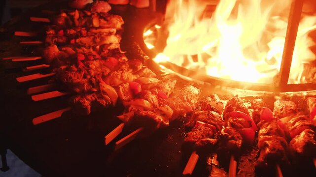 Close-up of meat on a skewer sizzling on a hot griddle beside an open fire. Flames and heat create an authentic outdoor cooking scene with rustic barbecue atmosphere.