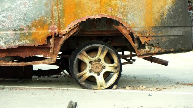 rusty automobile wheel rests against pavement beside heavily corroded vehicle frame. oxidised paintwork and decayed metal surfaces emphasize long term abandonment and dramatic structural damage.