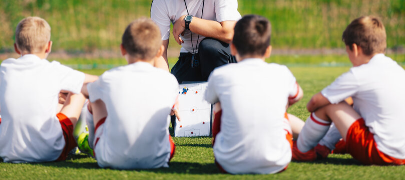 Youth Soccer Team in White Shirts Sitting on Grass Pitch Listening to Coach Holding Tactical Board - Pre-Match Briefing