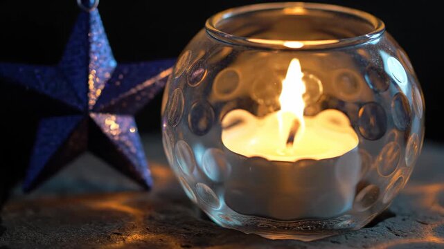 Small candle glows inside a round glass holder patterned with circular dimples, beside a blue glitter star on a dark rough surface, blending cool reflections and warm Christmas light in close focus