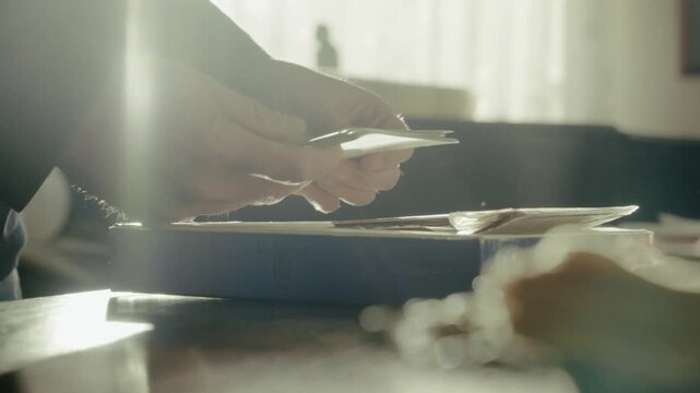 Person examines a box of old family photographs as warm sunlight filters through veiled curtains, creating a nostalgic and reflective atmosphere.