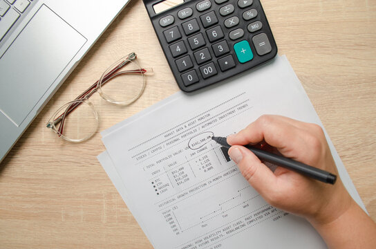 Top view of financial analyst checking investment portfolio report with pen, calculator, and laptop on wooden desk. Market data and asset monitoring concept.