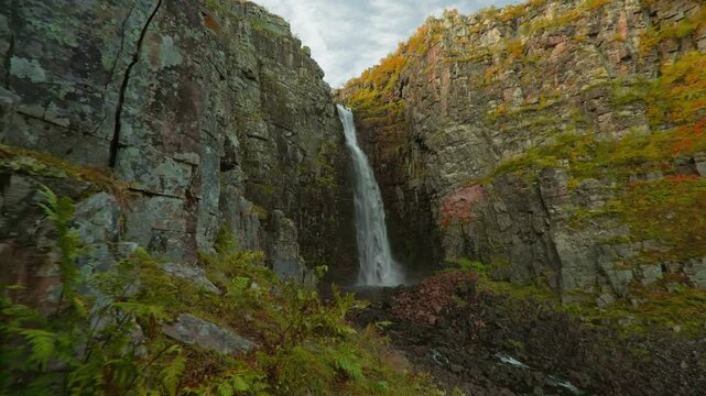 Cinematic push-in along a rugged canyon wall toward impressive Njupesk&auml;r Waterfall in Fulufj&auml;llet National Park Sweden, surrounded by golden autumn foliage with a blue sky and green fern leaves.