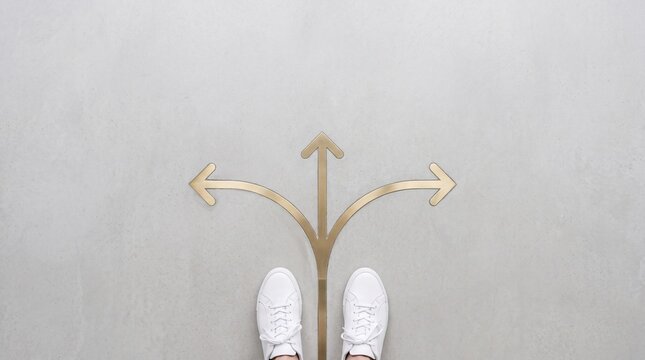 Overhead top view of person in white sneakers standing before three golden arrows pointing in different directions on a grey floor representing decision making and life choices.