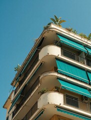 Fototapeta premium Low angle view of old apartment building with blue awnings. Mediterranean style architecture features white balconies, green plants on rooftop. Sunny day in European city with summer vibes. Urban
