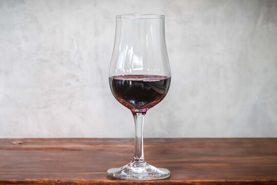 A glass of red wine on a dark wooden table against a textured grey concrete wall, with copy space &mdash; tulip-shaped tasting glass holding deep ruby wine in soft natural light.