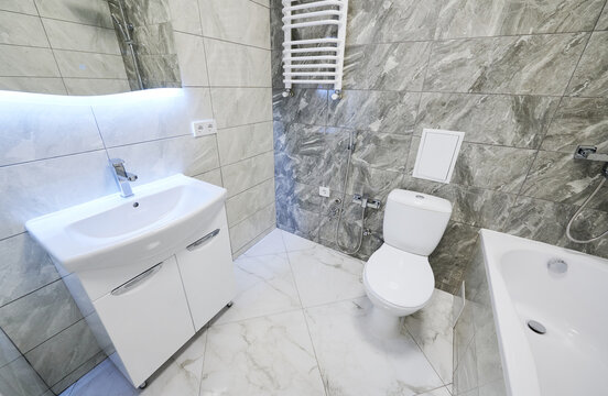 Fresh bathroom interior with sink vanity, toilet and bathtub in marble texture. Bright washroom with gray stone wall tiles, heated towel rail, glowing mirror and neat finish