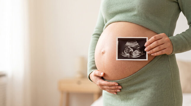 Pregnant woman holds ultrasound scan photo against her bare belly, embracing maternal joy