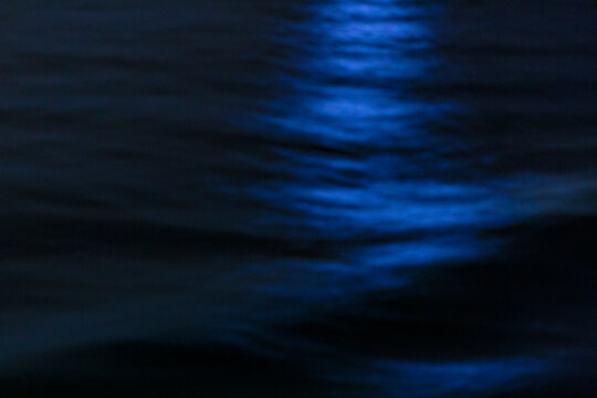 The ocean surface, midnight blue colours in the darkness, reflection, a ray of light on the water, and ripples.