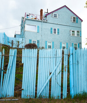 Low angle view of a broken fence, and a blue clapboard house with blue shutters. 