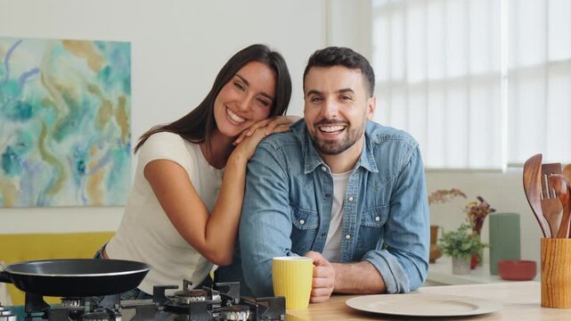 A smiling couple stands at a sunny kitchen island, resting on each other with casual warmth as they chat and laugh. A gas stove and decorative items frame the scene, creating a cozy, inviting family