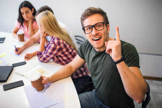 Cheerful nice young man in glasses looks on camera. He rocks. Other three young people sit behind hm and work in team.