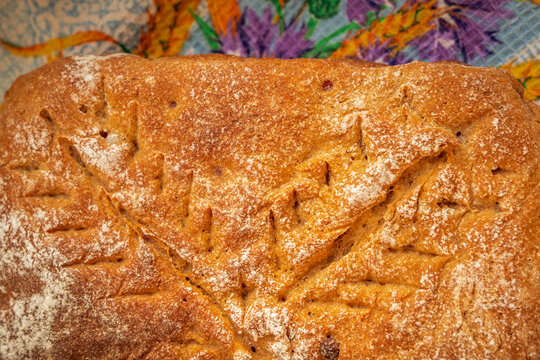 Homemade rectangular loaf with a rich golden-brown crust, decorative wheat-like scoring, light flour dusting, and scattered baked fruit pieces