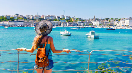 Fototapeta na wymiar Young female traveler with backpack and sun hat leaning on railing overlooking coastal harbor town with boats on sunny day