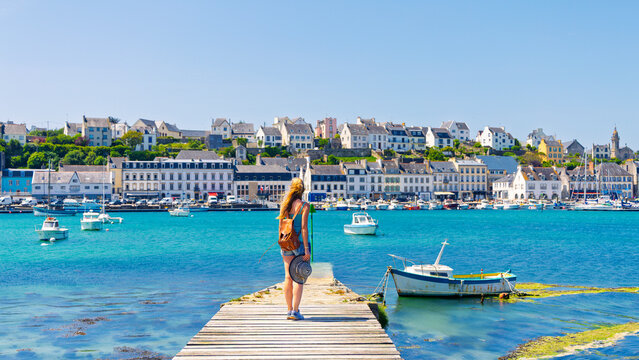 Young female traveler with backpack holding sun hat standing on wooden pier looking at coastal harbor town with boats on sunny day