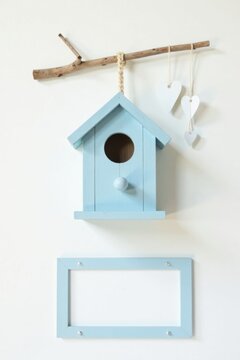 A charming pastel blue birdhouse hangs from a rustic branch, adorned with delicate heart decorations, complementing a matching pale blue frame on a pristine white wall.