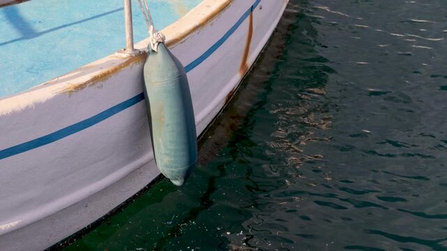 A boat rests on calm water with a fender hanging from its side. The scene shows clear blue water and a part of the boat's hull. It is a sunny day.