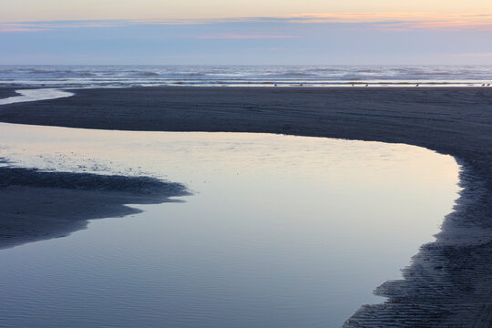 A curving stream flows across a beach into the ocean at dusk.