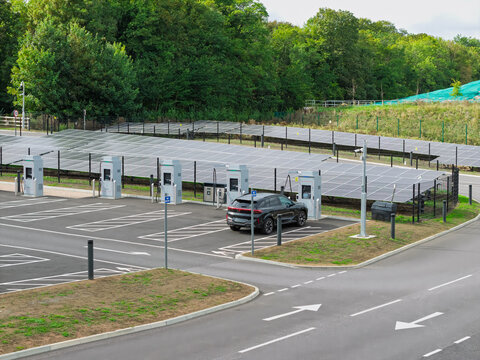 Aerial view of an electric vehicle charging station with a black SUV parked at a charger, featuring rows of solar panels and a paved parking lot in the United Kingdom.