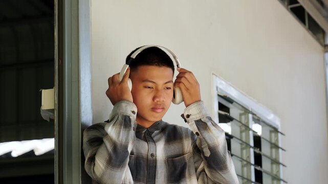 Asian teenage boy with buzz cut listening to music on white headphones, indoor portrait
