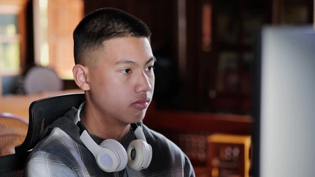 Asian teenage boy with buzz cut sitting at computer with white headphones around neck, side view