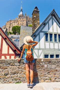 Vertical view of female traveler with straw hat looking at Mont Saint-Michel abbey and timber framed houses in Normandy France