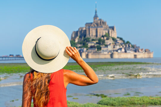 Young female traveler with straw hat looking at blurred Mont Saint-Michel abbey across tidal bay in Normandy France