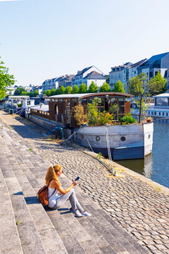 Young female listening music with earphones, sitting on stone stairs using smartphone near houseboats in Rennes Brittany France