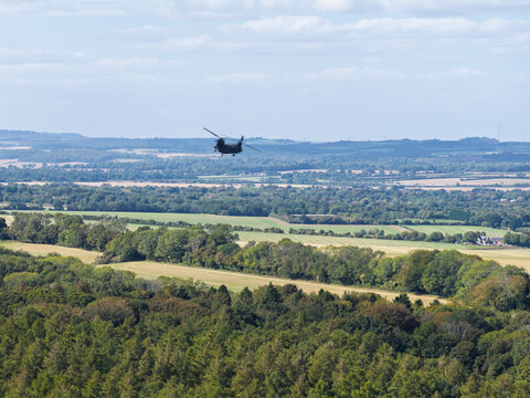 Aerial view of a military helicopter flying over a lush rural landscape with rolling green hills, dense forests, and agricultural fields in the United Kingdom.