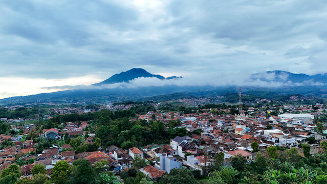 Aerial view of Tanjungsari town with dense residential rooftops, lush green trees, and a prominent mountain peak under a cloudy sky in Sumedang, West Java, Indonesia.