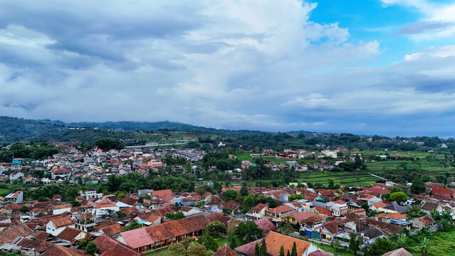 Aerial view of Tanjungsari featuring dense residential rooftops, lush green rice fields, and rolling hills under a cloudy sky in Sumedang, West Java, Indonesia.