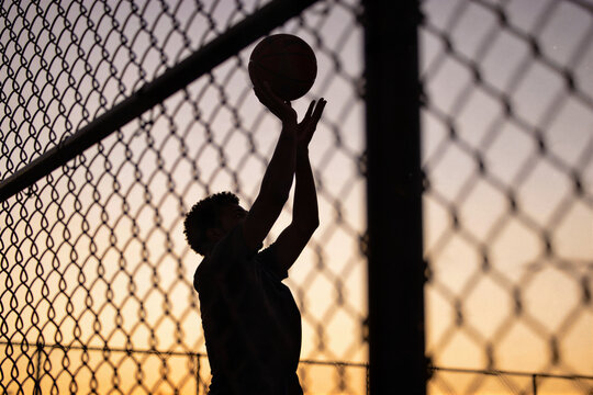 Basketball player shooting silhouette behind chain-link fence at golden hour