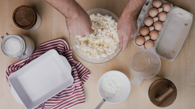 Top view man smashes cottage cheese with a fork for curd casserole.Fresh raw eggs, ceramic baking dish and dairy prepare a delicious, healthy and sweet family breakfast dessert.High quality 4k footage