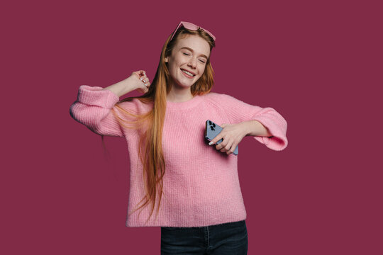 Smiling young woman holding smartphone with eyes closed in pink sweater and sunglasses on head against solid maroon studio background casual lifestyle portrait